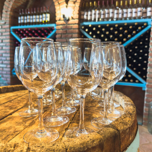 AdobeStock_1426149365 Many glasses of white wine on wooden barrel. Wine tasting event. Wine cellar with wine barrels. French wooden barrels. Wine shop wine bar winery or wine tasting concept